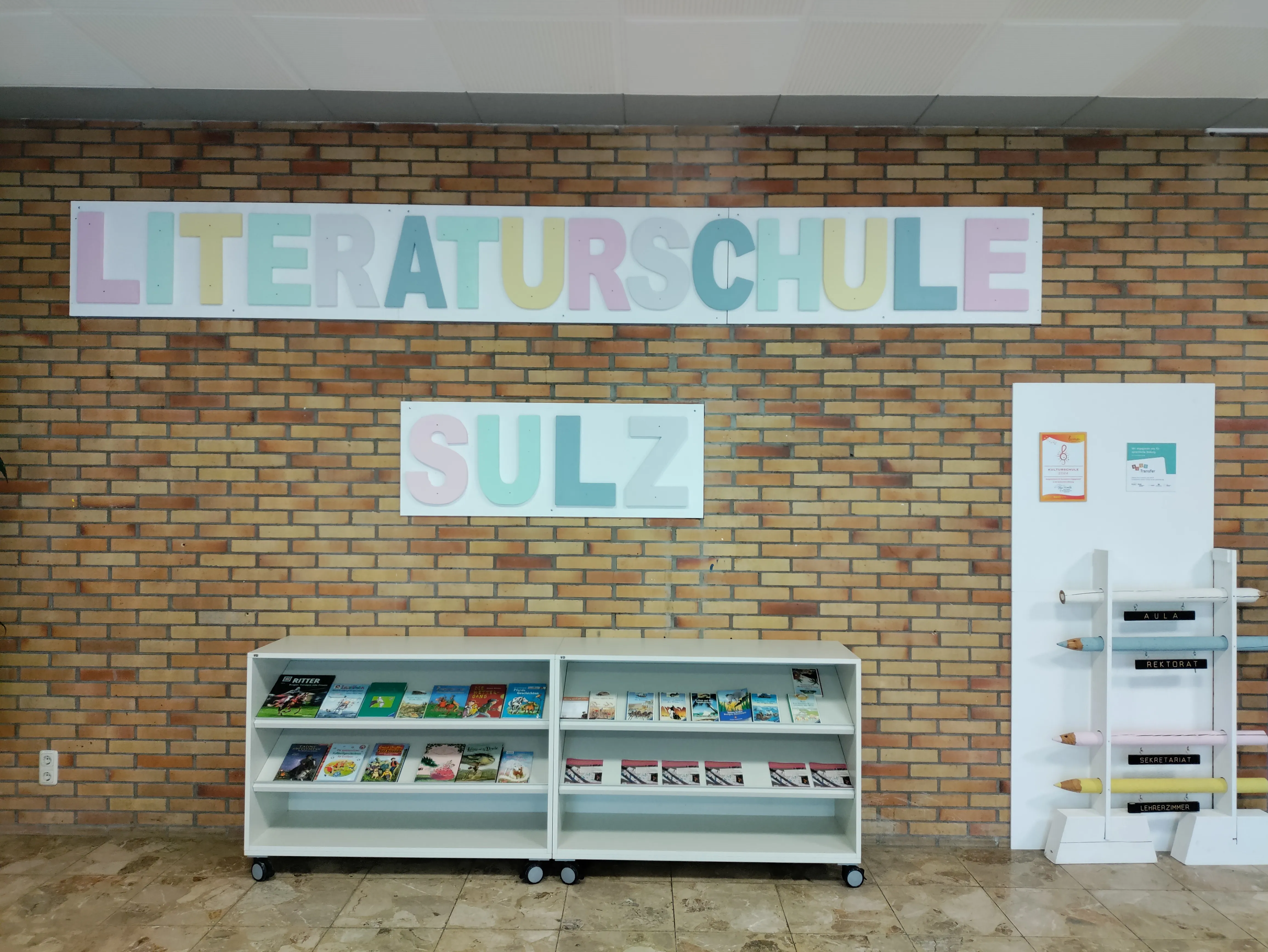 Backsteinwand im Foyer mit dem Schriftzug „Literaturschule Sulz“ aus bunten Buchstaben, darunter niedrige Regale mit ausgestellten Büchern.